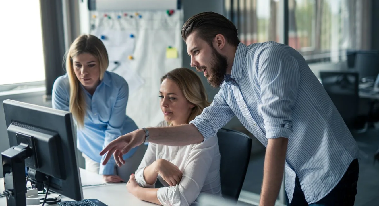 Three colleagues gathered around a monitor, one pointing at the screen