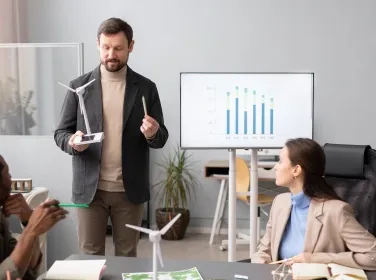 A presenter holding a wind-turbine model beside a bar chart on screen