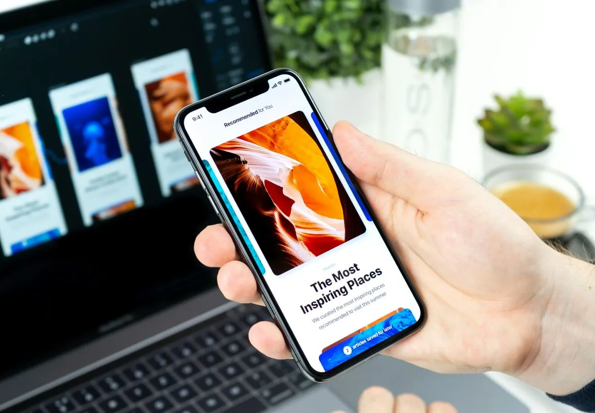 A hand holding a phone showing an article interface, with the same screens on a laptop behind it