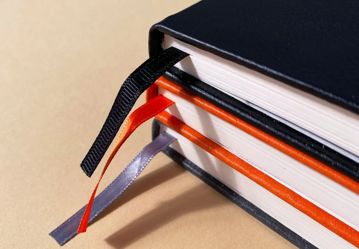 Stacked hardback books in black and orange with ribbon markers, on a sand-coloured background